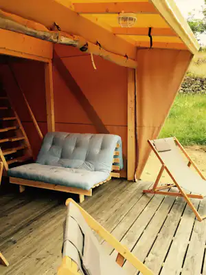 Offenes Zelt auf einer Holzterrasse mit gepolstertem Sofa und Liegestuhl, seitlich sichtbarem Holzrahmen und Vorhang.
