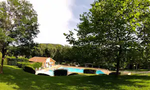 Campingplatz mit Pool im Grünen, umgeben von Bäumen und Hecken, im Hintergrund ein Gebäude mit Ziegeldach.