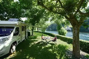 Wohnmobil am Flussufer im Grünen, zwei Campingstühle am Tisch unter Bäumen im Schatten.