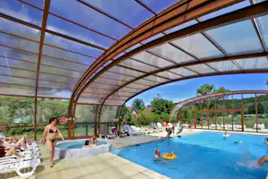 Überdachtes Camping-Schwimmbecken mit Liegestühlen, Kindern im Wasser und Bäumen im Hintergrund.