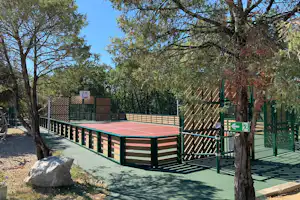 Tennisplatz mit Umzäunung und Netz, gepflasterter Weg am Rand, Bäume und grüner Zaun im Hintergrund.