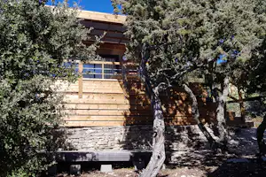 Holzterrasse mit Geländer vor einem Gebäude, umgeben von Olivenbäumen und Hecken am Campingplatz.