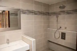 Moderne Dusche mit Regendusche, Fliesenwand und Badewannenrand, Fenster mit Holzjalousien im Hintergrund.