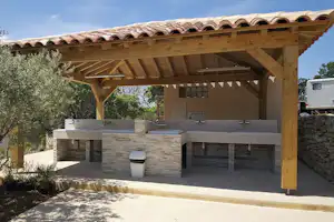 Überdachter Grillbereich mit Holzpergola, Edelstahlgrills und Spülbecken, Steinmauer und Abfalleimer auf dem Campingplatz.