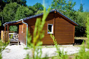 Holzhütte mit Veranda, Fenster und Regenrohr, umgeben von Bäumen und Gras, im Vordergrund Kiesweg.