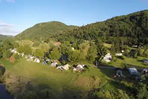 Campingplatz mit Zeltwiese, Wohnwagen und Autos, umgeben von Bäumen und Hügeln im Hintergrund.