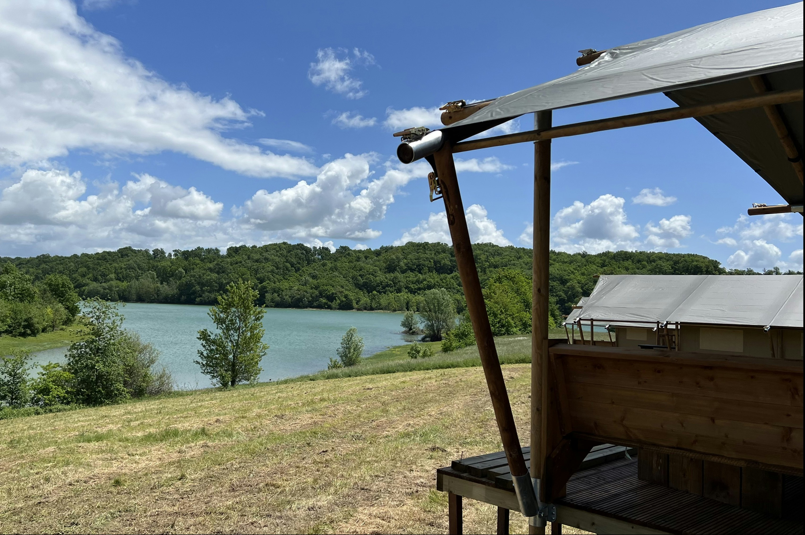 Blick vom Campingunterstand auf einen türkisfarbenen See, Wiese und Bäume unter blauem Himmel mit Wolken.