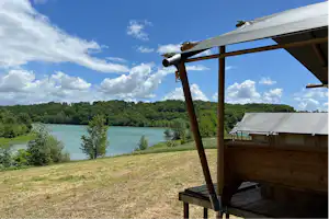 Blick vom Campingunterstand auf einen türkisfarbenen See, Wiese und Bäume unter blauem Himmel mit Wolken.