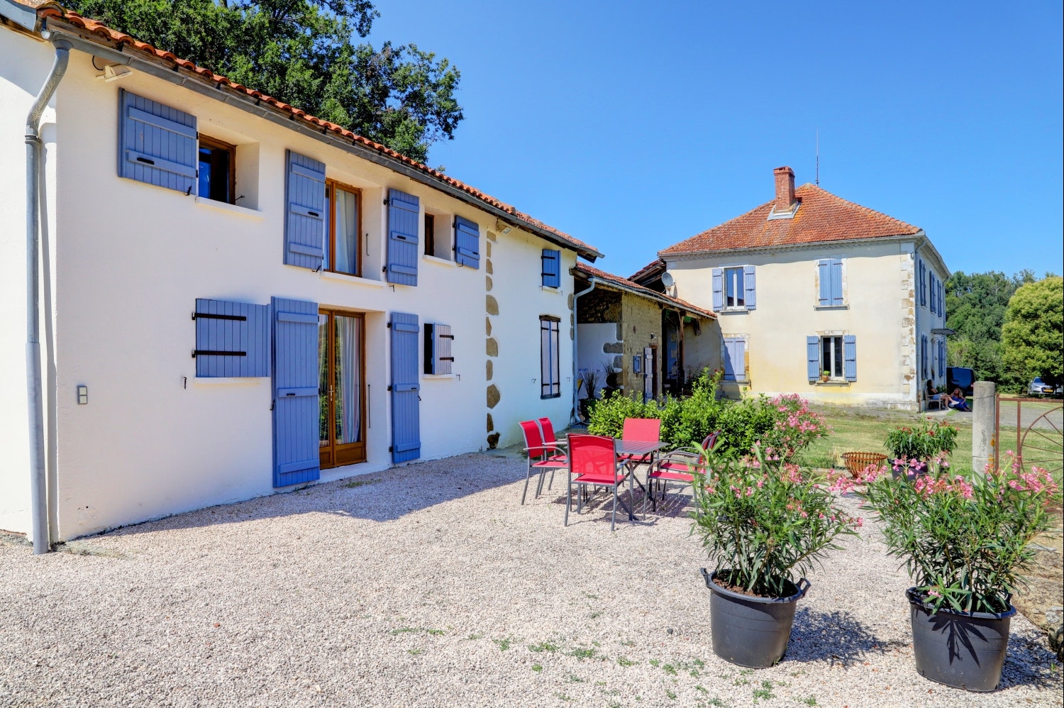 Ferienhaus mit blauen Fensterläden, Kiesvorplatz und Garten mit Blumen in Töpfen vor zwei Gebäudeteilen.