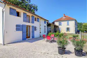 Ferienhaus mit blauen Fensterläden, Kiesvorplatz und Garten mit Blumen in Töpfen vor zwei Gebäudeteilen.