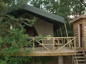 Camping-Unterkunft auf Stelzen mit Veranda, Holzgeländer und Markise, umgeben von Büschen und Bäumen.