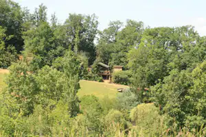 Blick auf einen Campingplatz am Hang mit Bäumen und Wiesen, im Hintergrund einzelne Gebäude und Wege.