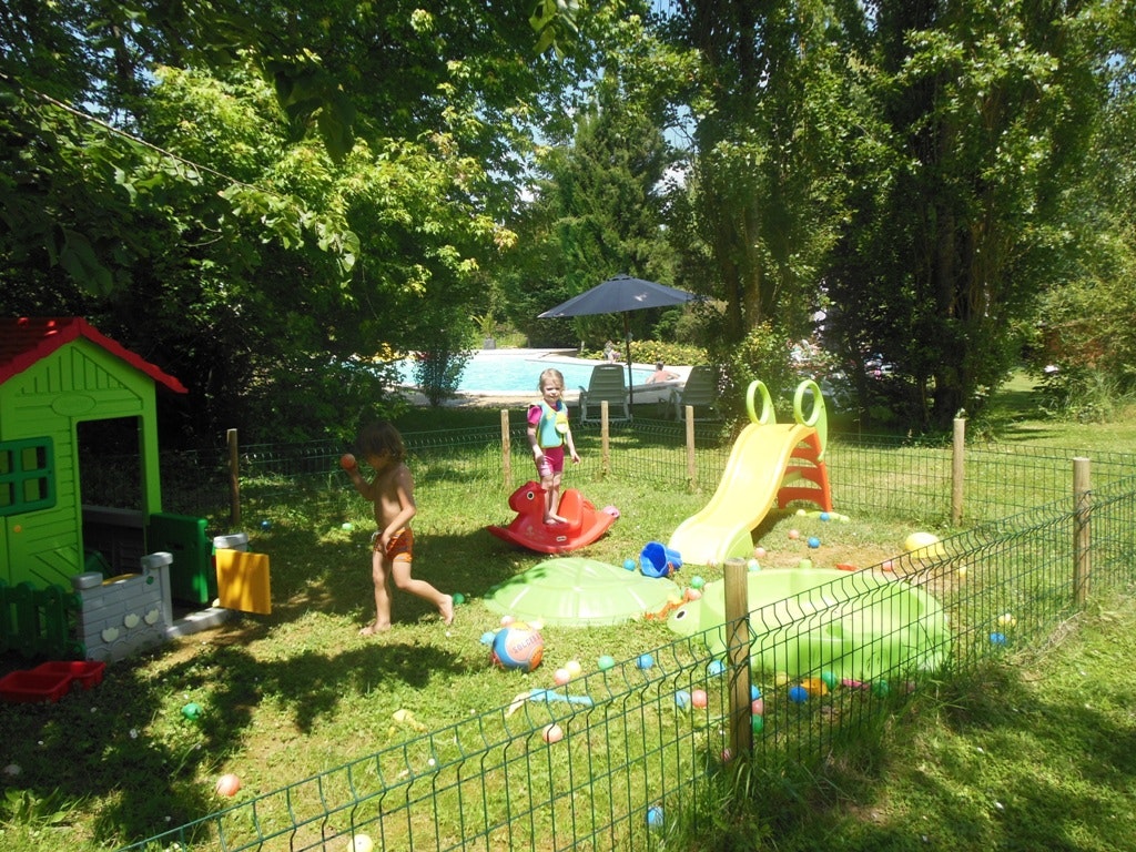Spielbereich im Grünen: Kinder spielen im Planschbecken mit Rutsche, daneben Zäune, Spielzeug und ein grünes Spielhaus.