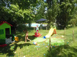 Spielbereich im Grünen: Kinder spielen im Planschbecken mit Rutsche, daneben Zäune, Spielzeug und ein grünes Spielhaus.