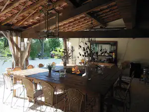 Überdachte Terrasse mit Holztisch, Stühlen und Feuerstelle im Kaminbereich, daneben Pflanzen und Grillutensilien an der Wand.
