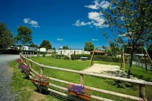 Campingweg mit Holzgeländer, Blumenbeeten und Rasenfläche; dahinter stehen Wohnwagen und mobile Häuser zwischen Bäumen.