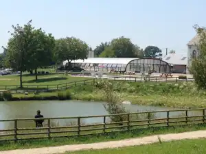 Campingplatz mit Teich, Holzgeländer und Kiesweg; im Hintergrund ein Gewächshaus und Gebäude neben Bäumen.
