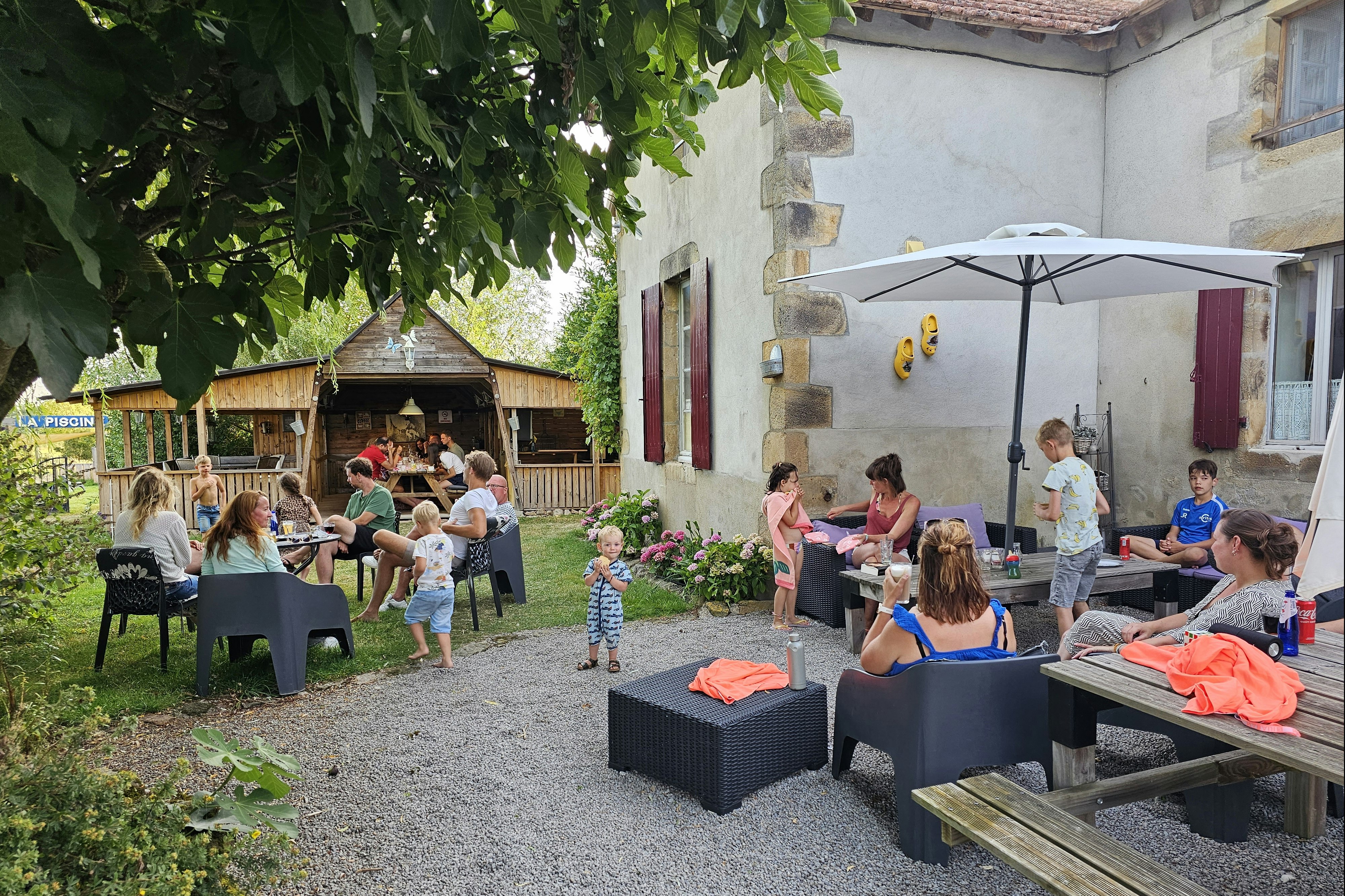 Familie sitzt im Campinggarten an einem Tisch unter Sonnenschirm, mit Kindern, Blumenbeeten und Holzunterstand im Hintergrund.