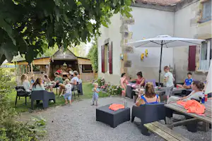 Familie sitzt im Campinggarten an einem Tisch unter Sonnenschirm, mit Kindern, Blumenbeeten und Holzunterstand im Hintergrund.