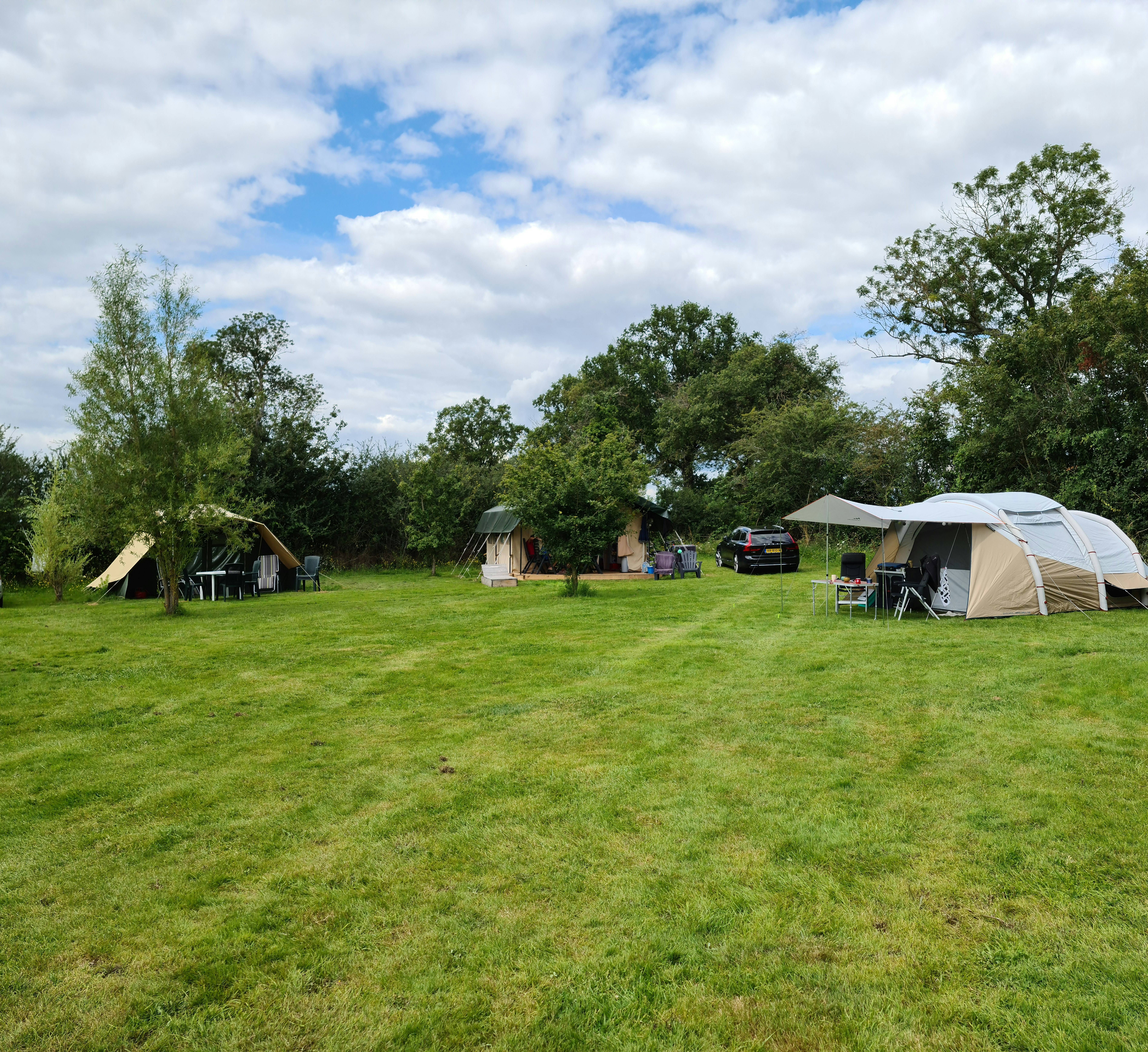 Campingplatz mit Zelten, Sitzgarnitur und Tisch auf grünem Rasen, umgeben von Bäumen und einem schwarzen Auto.