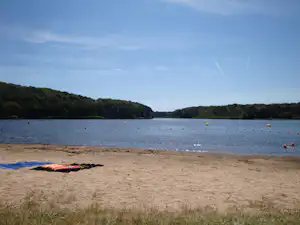Sandstrand am See mit Liege und Blick auf das ruhige Wasser, im Hintergrund bewaldete Hügel und Bojen.