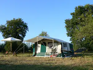 Campingzelt mit Vorzelt, Stühlen und Sonnenschirm auf einer Wiese, im Hintergrund Bäume und blauer Himmel.