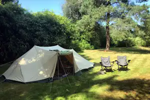 Campingzelt auf einer grünen Wiese im Garten, daneben zwei Klappstühle und ein Tisch, umgeben von Bäumen und Hecken.