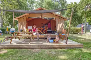 Familienzelt auf einer Holzplattform mit Markise, Campingstühlen und Tisch im Grünen zwischen Bäumen.
