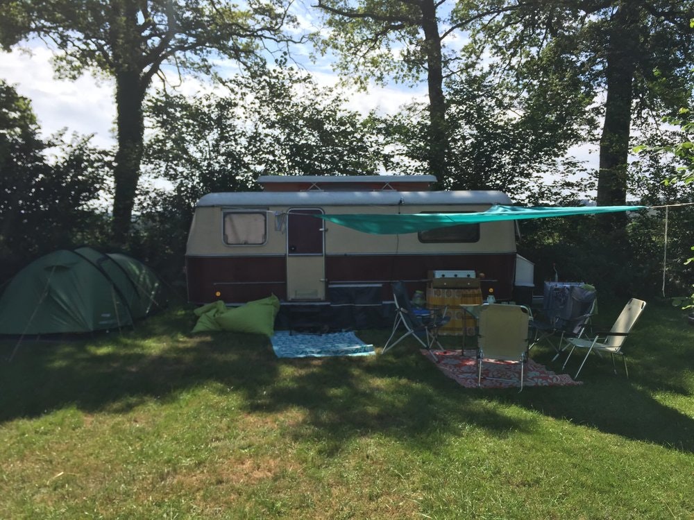 Wohnwagen auf einem Campingplatz im Grünen, mit Vorzelt, Gartenmöbeln und Sonnenschirm zwischen Bäumen.