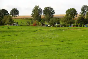 Campingwiese mit mehreren Zelten, Bäumen und Büschen, im Hintergrund Felder und ein kleiner Wohnwagen.