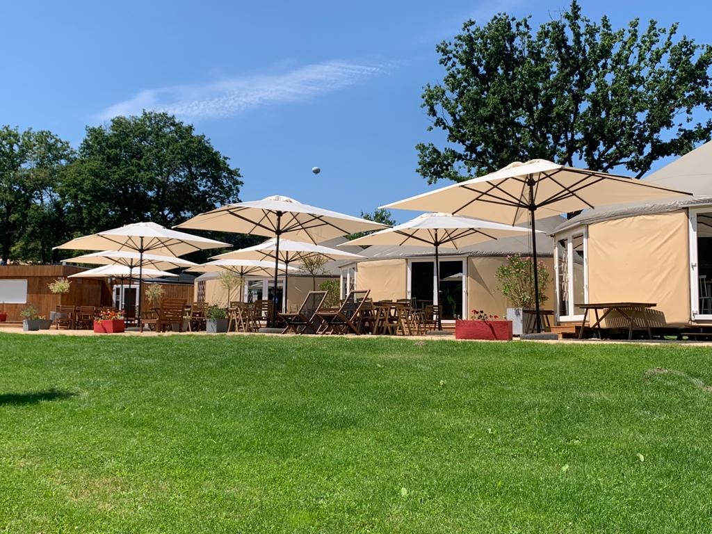 Terrasse am Campingplatz mit mehreren Sonnenschirmen, Tischen und Stühlen vor weißen Zelthäusern, umgeben von Rasen und Bäumen.