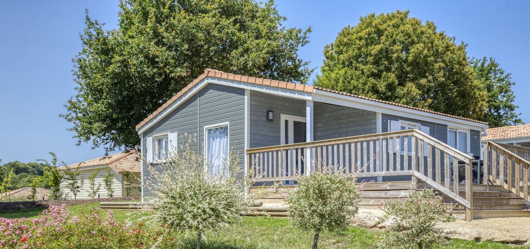 Holz-Ferienhaus mit Veranda und Geländer, davor Sträucher und ein gepflasterter Weg im Campingbereich.