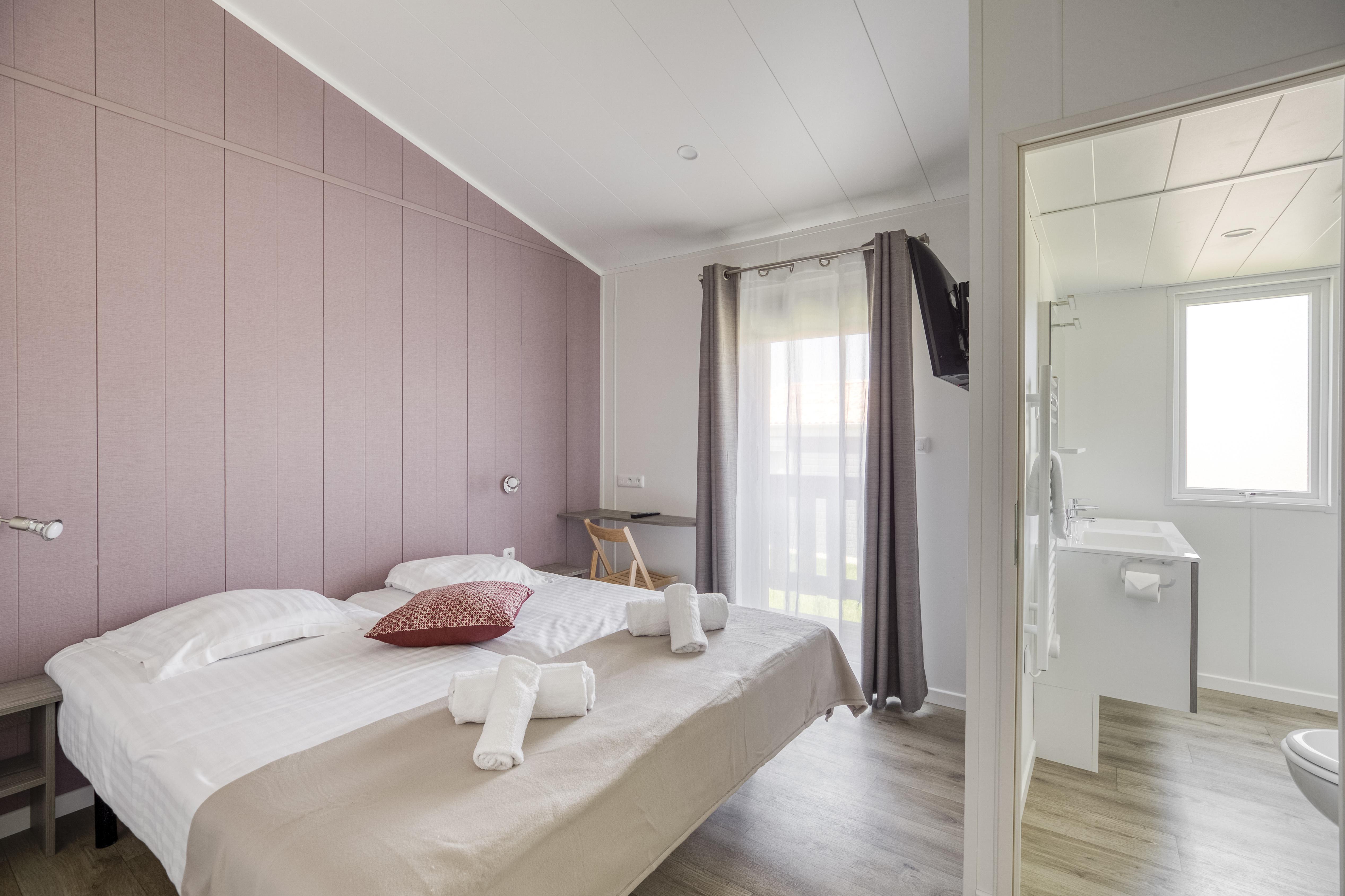 Schlafzimmer mit Doppelbett, rosa Wand, Nachttisch und Vorhängen; Blick ins angrenzende Badezimmer mit Dusche und WC.