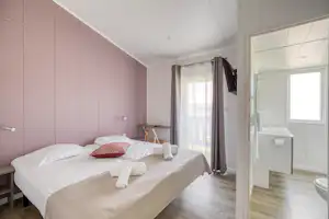 Schlafzimmer mit Doppelbett, rosa Wand, Nachttisch und Vorhängen; Blick ins angrenzende Badezimmer mit Dusche und WC.