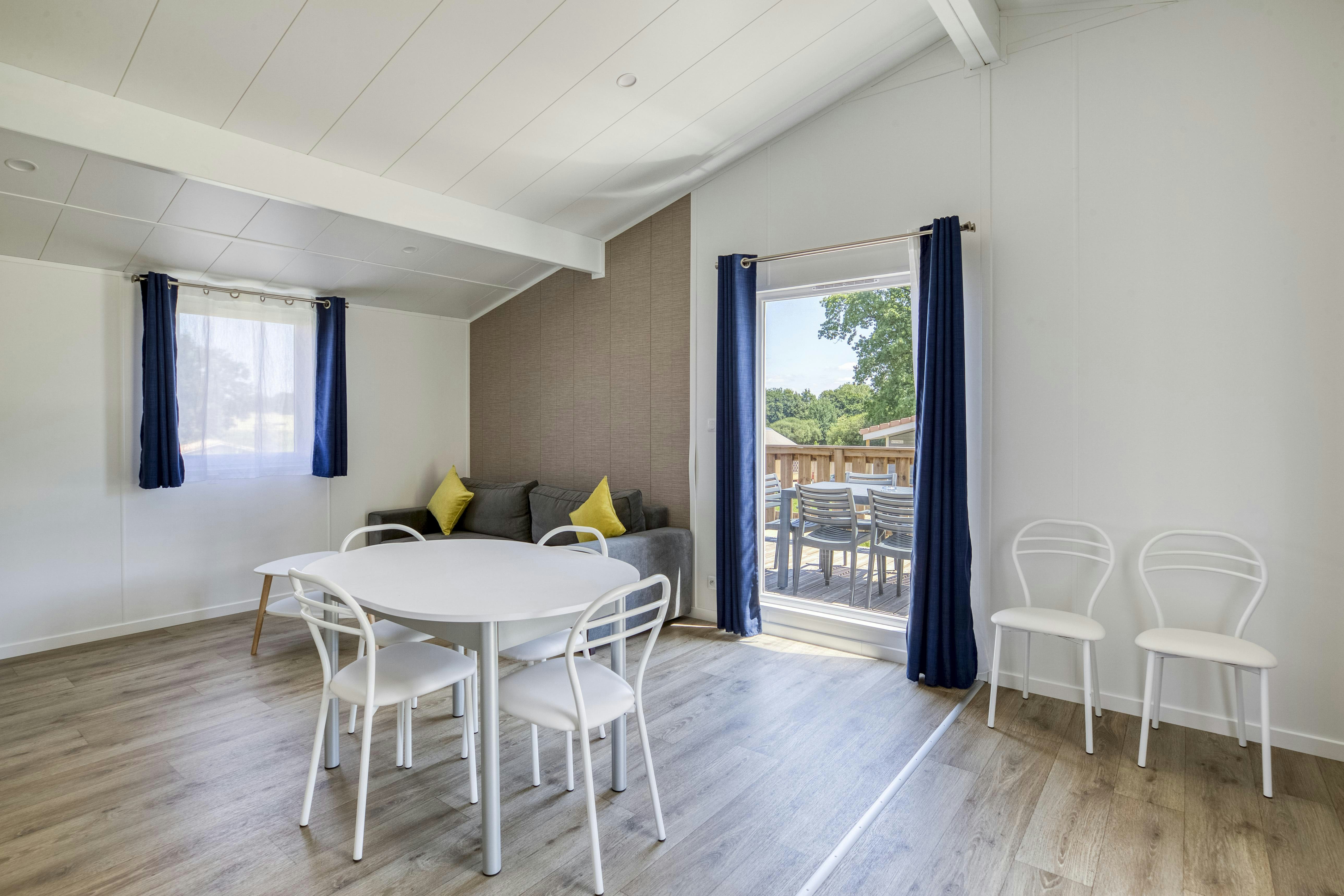 Wohnbereich mit Esstisch und weißen Stühlen, Sofa mit Kissen und Zugang zur Terrasse durch große Schiebetür