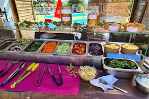 Frühstücksbuffet mit Schüsseln voller Kräuter, Gewürze und Gemüse, daneben Besteck auf einem Tisch im Campingbereich.