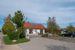 Campinggebäude mit rotem Dach, gepflastertem Weg und Parkplatz; davor Bäume und ein schwarzes Auto.