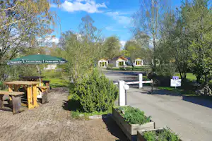 Campingplatz mit gepflastertem Weg, Heckenbeeten und mehreren kleinen Häusern, umgeben von Bäumen und blauem Himmel.