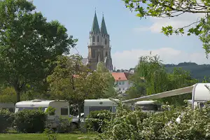 Wohnwagen auf dem Campingplatz mit Blick auf eine Kirchturmspitze zwischen Bäumen und Büschen.