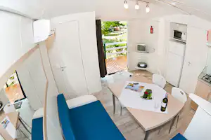 Innenansicht eines Wohnwagens mit Esstisch, Stühlen, Küche und Blick auf die Terrasse mit Garten im Hintergrund.