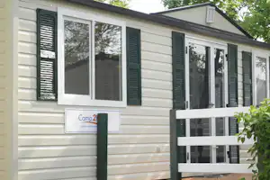 Mobiles Wohnheim mit großen Fenstern und grünen Rollläden, Schild „Camp2“ an der Hauswand, Hecke im Vordergrund.