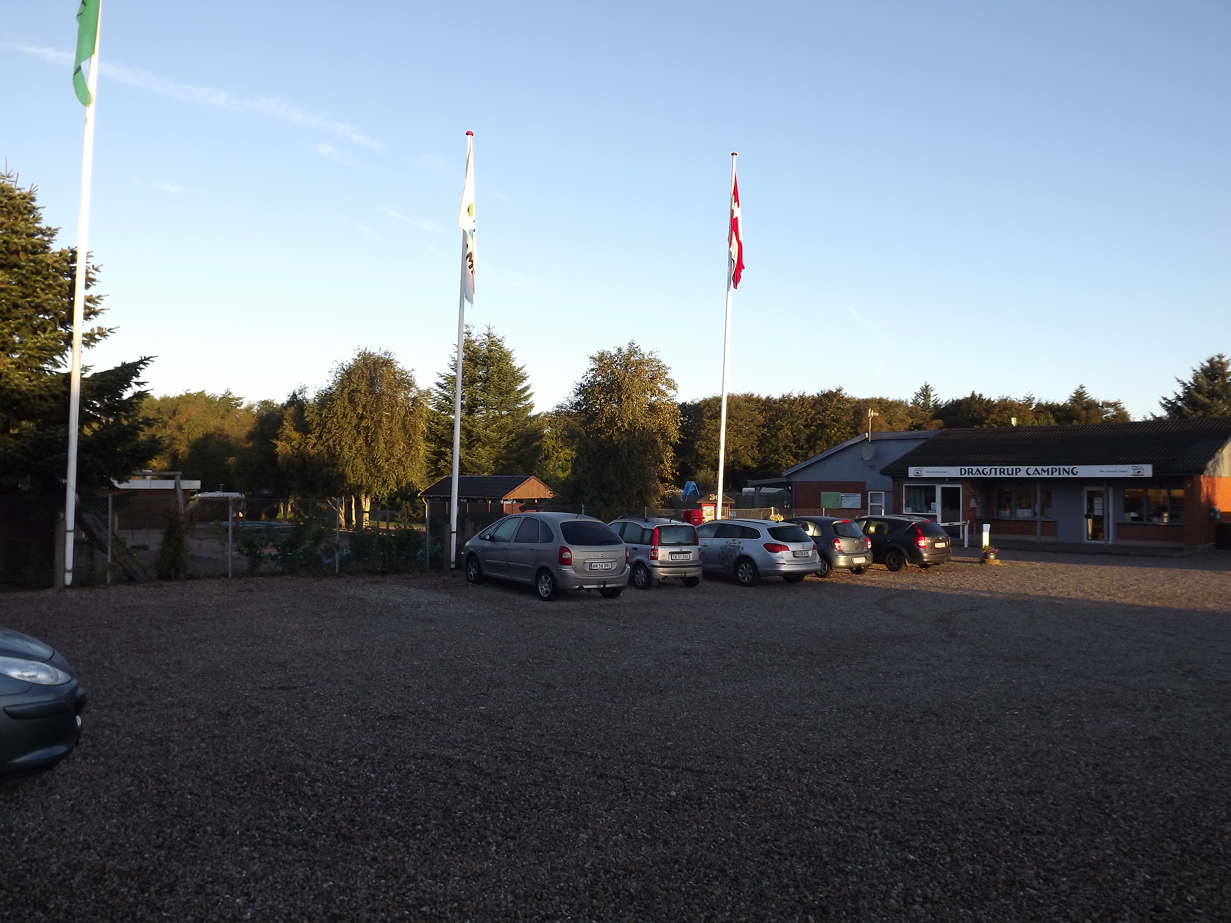 Campingplatz-Eingang mit Parkplatz, mehreren Autos und Fahnenmasten vor einem Gebäude am Rand des Geländes.