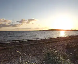 Strand mit Wellen und Blick aufs Meer bei Sonnenuntergang, im Hintergrund Wolken und eine dunkle Küstenlinie.