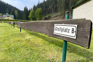 Schild „Stellplatz B“ an einem grünen Pfosten, daneben Wiese und Wald mit Nadelbäumen am Campingplatz.