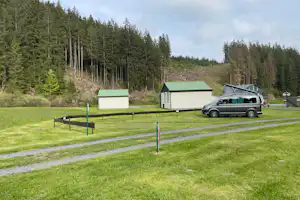 Grüne Campingwiese mit zwei Mobilheimen, Parkplatz und Auto am Rand, umgeben von hohen Nadelbäumen im Wald.