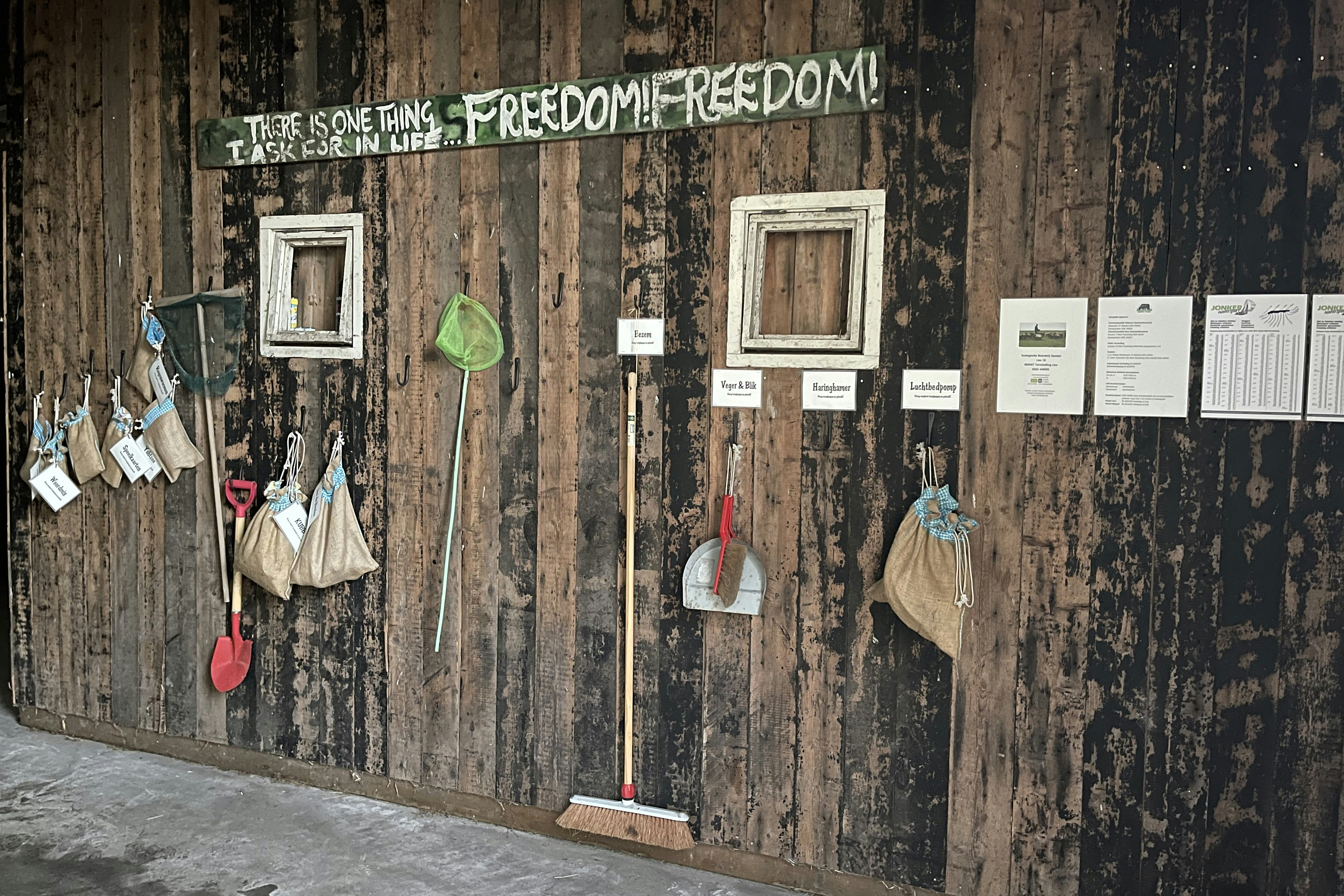 Holzwand mit mehreren hängenden Schildern und Zetteln, darunter ein grünes Herz und rote Haken, daneben ein Weg am Boden.