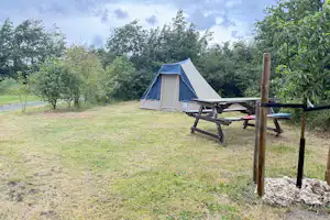 Zelt auf einem Campingplatz mit Picknicktisch und Bank, umgeben von Wiesen und Bäumen im Hintergrund.