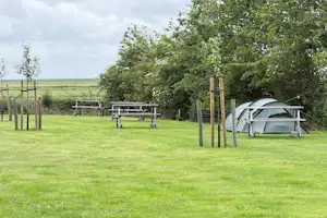 Campingzelt im Garten auf einem grünen Rasen, mit Picknicktisch und Sitzbank unter Bäumen am Zaun.