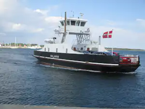 Fahrgastschiff mit dänischer Flagge, auf dem Wasser vor dem Ufer mit Segelbooten und Bäumen.