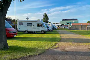 Reihe von Wohnmobilen auf einem Campingplatz-Stellplatz, mit Grasfläche, Schotterweg und Gebäuden im Hintergrund.